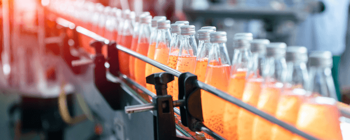 La imagen muestra una línea de embotellado con botellas de bebida de color naranja en un entorno industrial.