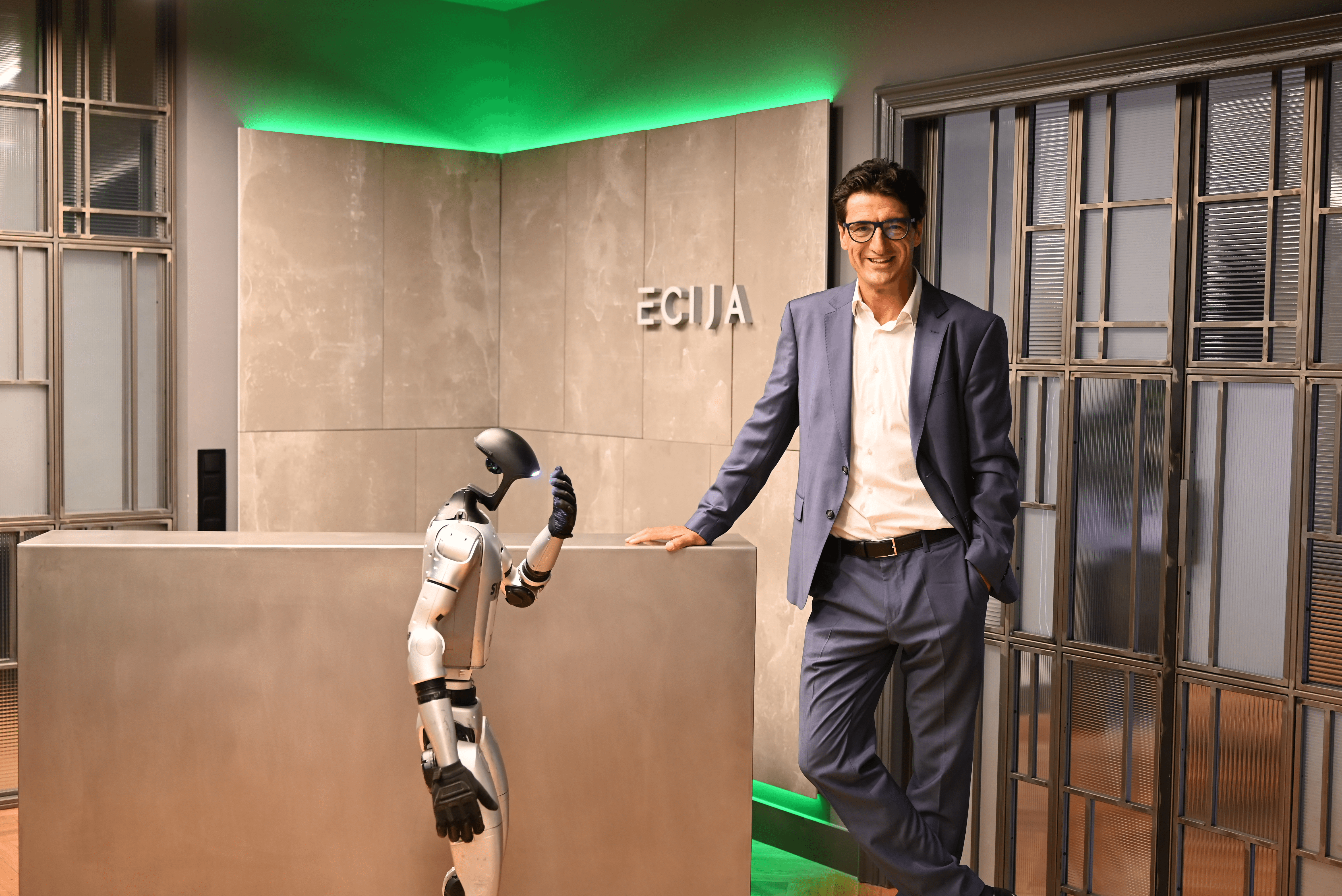 Un hombre sonriente vestido con traje está de pie junto a un robot humanoide en una oficina moderna.