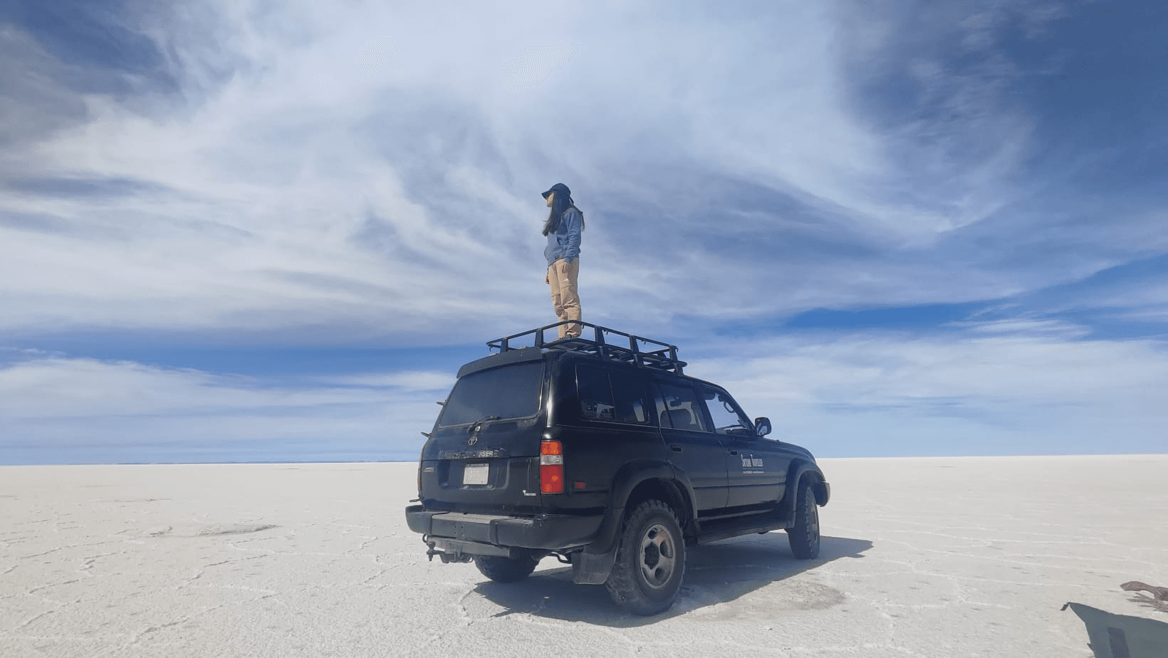 Una persona está de pie sobre el techo de un vehículo en un vasto paisaje de sal con cielo azul y nubes.