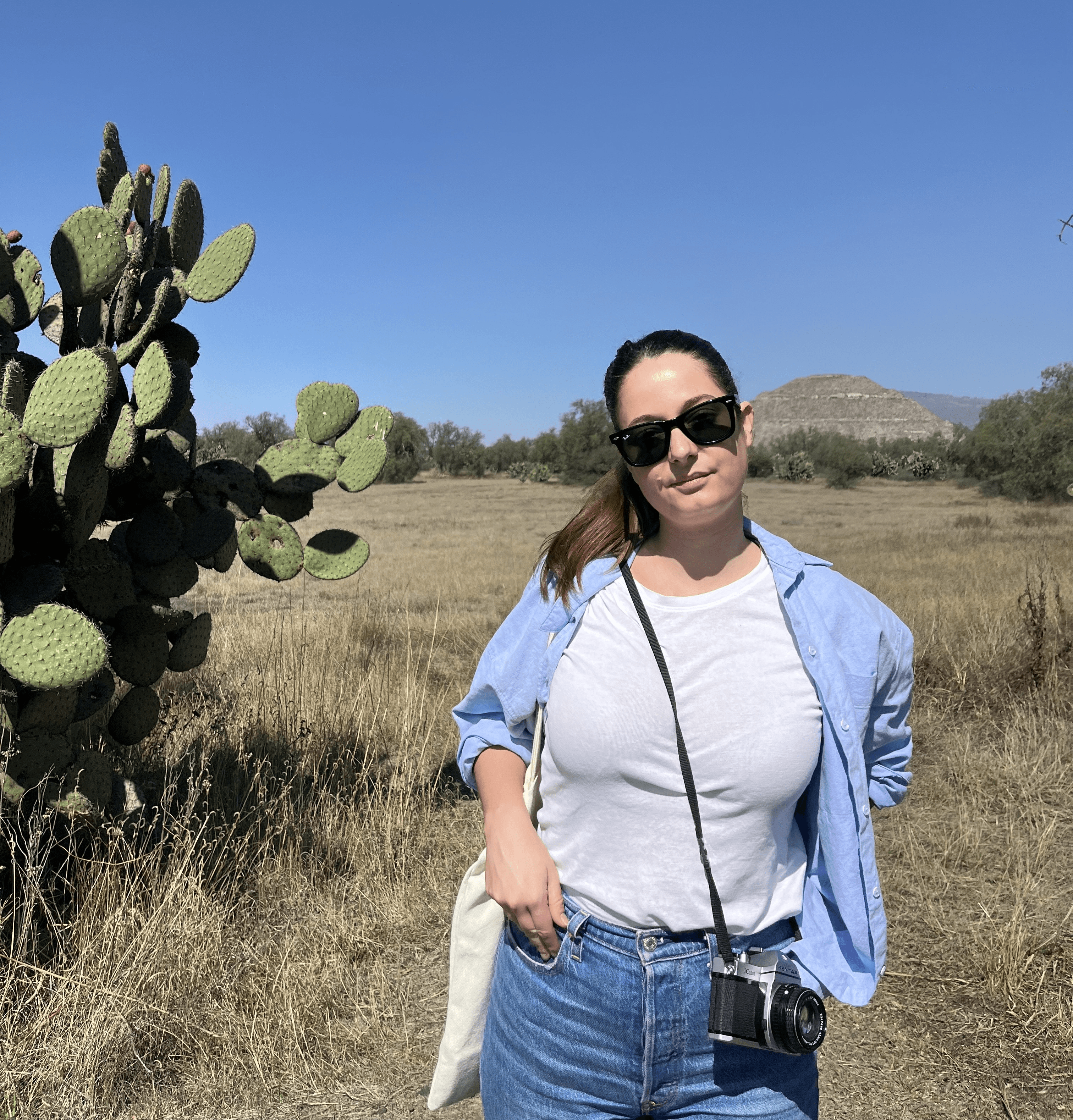 Una mujer sonriente con gafas de sol posa en un campo amplio junto a un cactus.