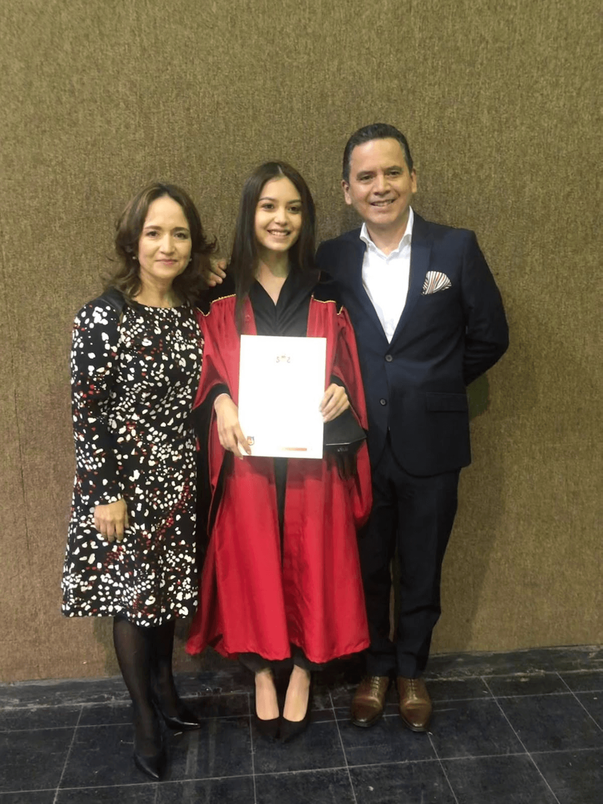 Tres personas posan juntas, una joven con toga y birrete sostiene un diploma entre un hombre y una mujer sonrientes.