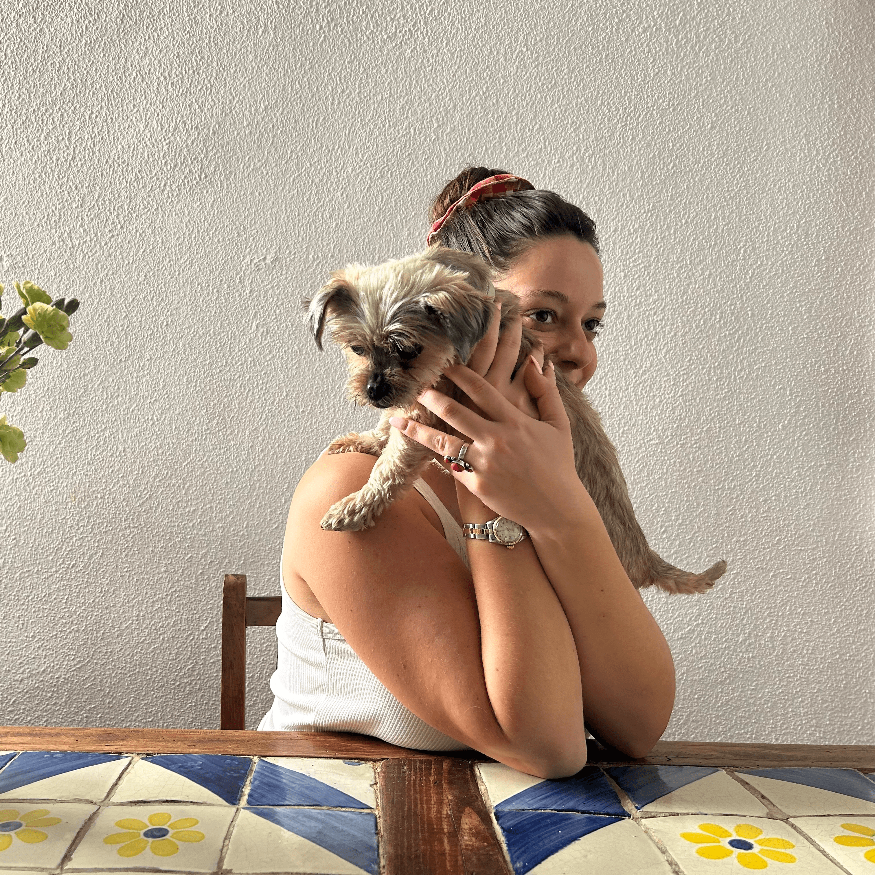 Una mujer sonriente sostiene un perro en brazos mientras está sentada en una mesa.